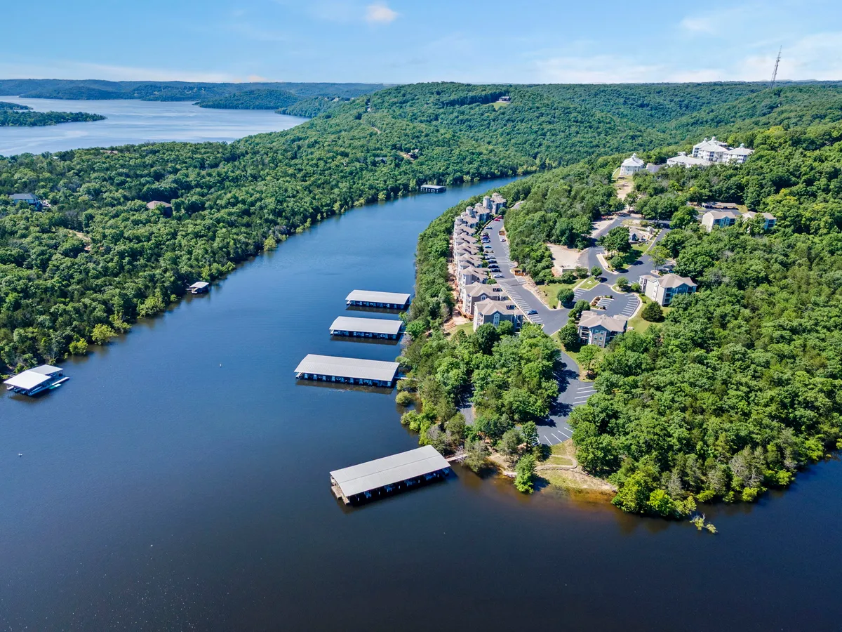 A tranquil, wide-angle view of the lake at sunset, with boathouses along the water's edge, green hills in the background, and a few residential homes visible. The calm river reflects golden hues, evoking a peaceful and inviting atmosphere for outdoor activities like boating, fishing, and lakeside picnics.