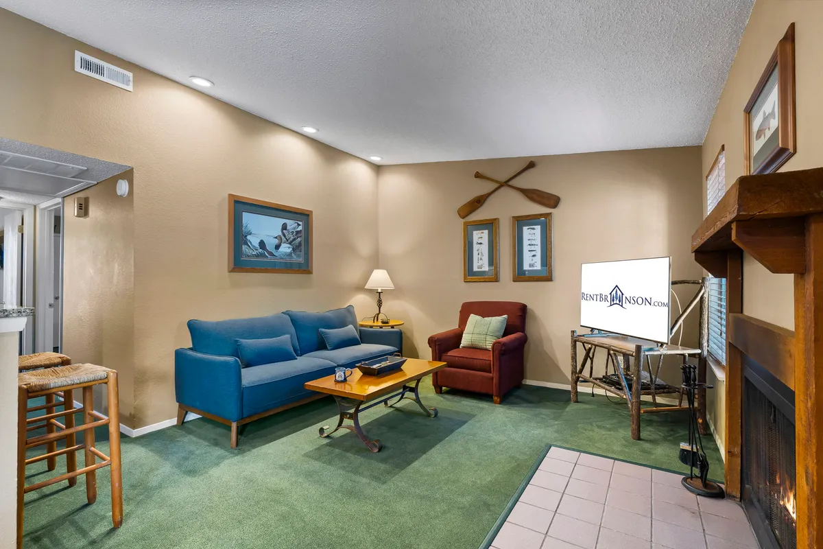 The image showcases a warm and inviting living area featuring a plush sofa, a cozy armchair, a stylish coffee table, a modern television, a crackling fireplace, and charming wall decor. The soft lighting and thoughtful layout create a perfect space for relaxation, family gatherings, or unwinding after a day at the lake.