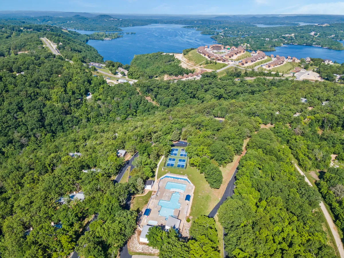 A serene resort exterior featuring the swimming pool in the foreground, surrounded by lush green landscaping, with a stunning lake view and residential buildings in the distance. The scene conveys a sense of calm, relaxation, and community.
