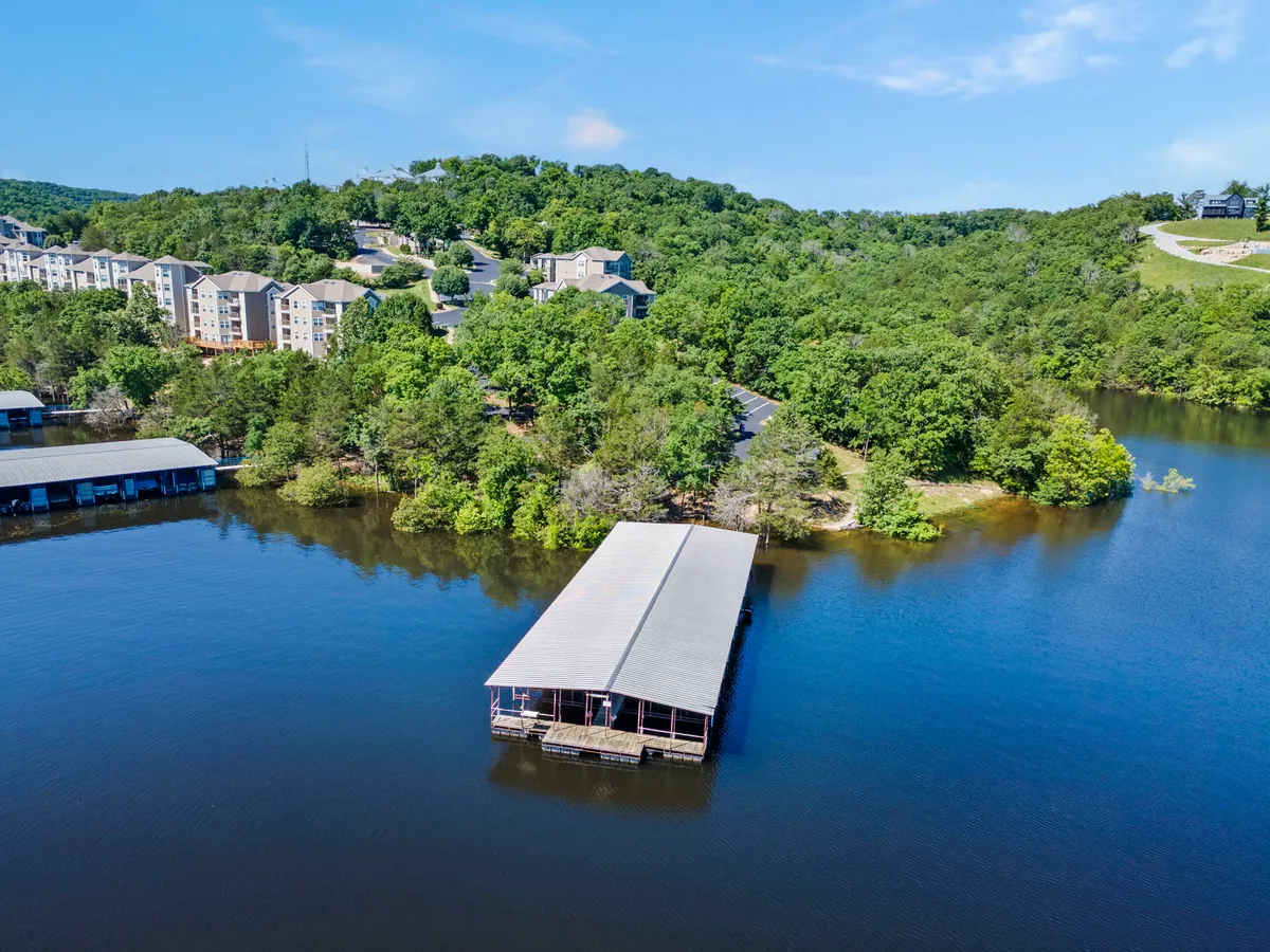 A wide exterior view captures the vibrant lake, boathouse, lush surrounding greenery, and residential buildings nestled along the shoreline. The scene is peaceful yet lively, suggesting possibilities for fishing, boating, and lakeside picnics in a scenic setting.