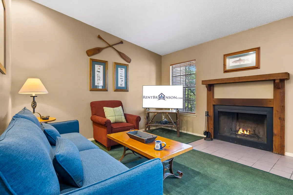 This image features a cozy living room with a sofa, fireplace, armchair, and coffee table. Warm lighting and the flicker of a wood-burning fireplace create a welcoming and comfortable environment, ideal for relaxing, reading, or socializing. Large windows allow for natural light and glimpses of the lush surroundings beyond.