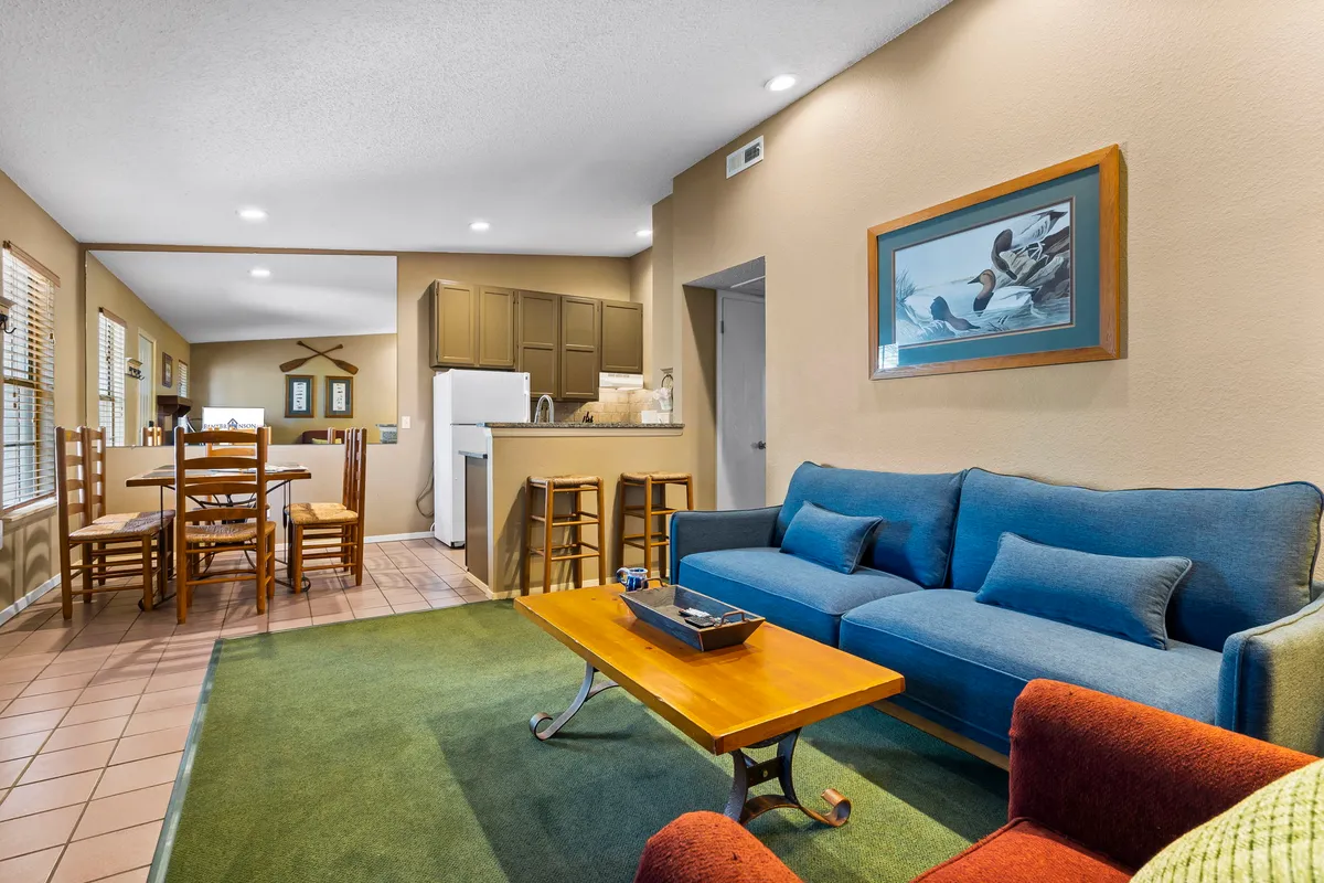 This wide-angle interior image displays a blue sofa, wooden coffee table, adjacent dining area, and kitchen counter, offering a broader perspective of the central living space. The setting radiates warmth and hospitality, ideal for both entertaining and relaxing.