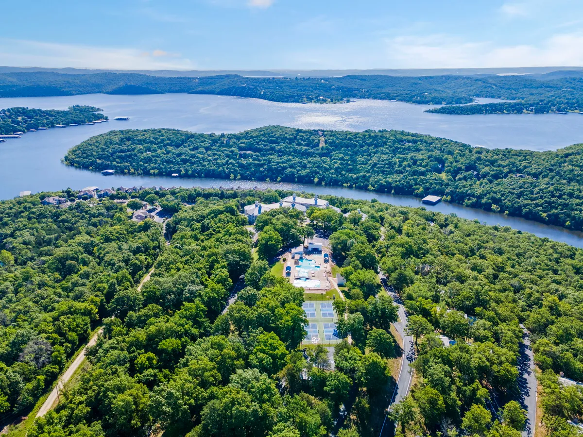 This exterior scene captures a sweeping view of the resort's outdoor swimming pool, nestled among lush greenery, with the sparkling lake and tennis court visible in the background. Residential homes dot the landscape, reinforcing the sense of a vibrant, activity-filled environment.