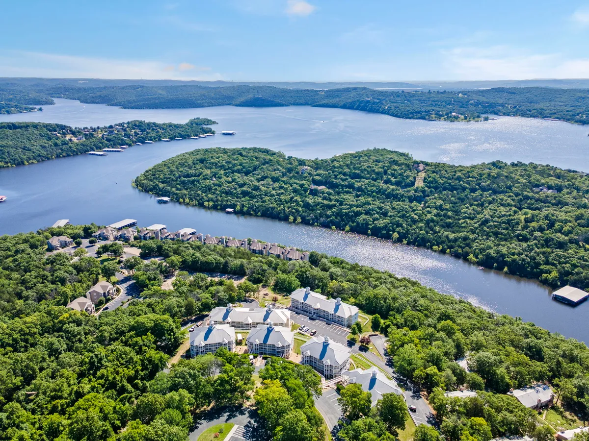 A wide, tranquil exterior view over Table Rock Lake, framed by lush greenery. The image captures residential buildings nestled along a curved shoreline, with expansive water views dominating the scene. The sun casts a soft glow, suggesting early morning or approaching evening, ideal for romantic walks or quiet reflection.